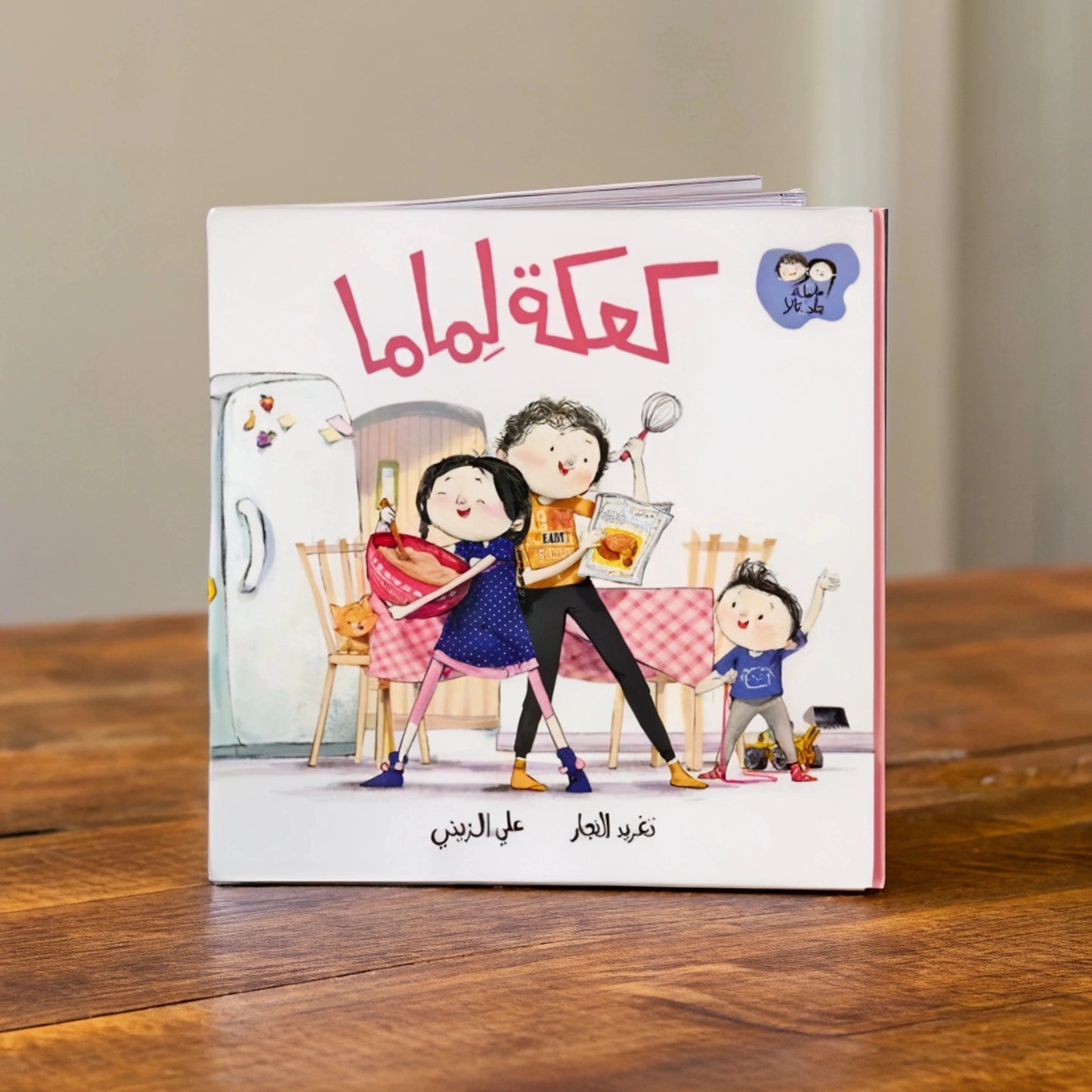 Illustration from the Arabic children's book 'Cake for Mama,' showing three happy siblings baking a cake as a surprise for their mother. The eldest reads a recipe, the middle child stirs the batter, and the youngest cheers excitedly. A cat watches nearby, adding warmth to the scene.