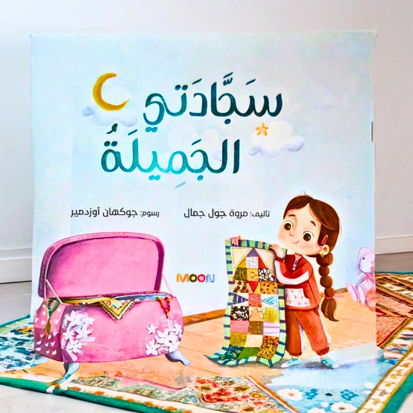 Sajadati Al Jameela Arabic children's book displayed on a beautifully patterned prayer mat. The book cover features an illustrated girl holding a colorful patchwork prayer mat, emphasizing the story’s theme of faith and prayer for young Muslim kids.